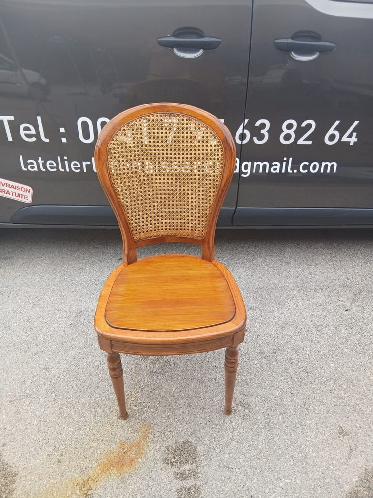 Wooden chair with warm honey-colored rattan woven backrest and polished wooden seat displayed on asphalt in front of workshop van. Van displays contact information Tel: 079 563 82 64 and latelierdelarenaissance@gmail.com. Chair shows professional craftsmanship with intact caning in tight crosshatch pattern and clean wood finish. Setting conveys completed restoration work with proud professional presentation of finished furniture piece