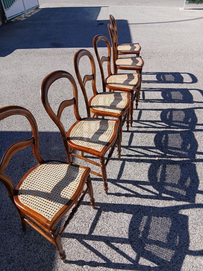 Wooden chair with warm honey-colored rattan woven backrest and polished wooden seat displayed on asphalt in front of workshop van. Van displays contact information Tel: 079 563 82 64 and latelierdelarenaissance@gmail.com. Chair shows professional craftsmanship with intact caning in tight crosshatch pattern and clean wood finish. Setting conveys completed restoration work with proud professional presentation of finished furniture piece